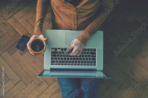 Fotografía Portátil y una taza de café en las manos de la muchacha que se sienta en un suel