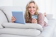 © WavebreakMediaMicro - Smiling blonde lying on couch shopping online with tablet
