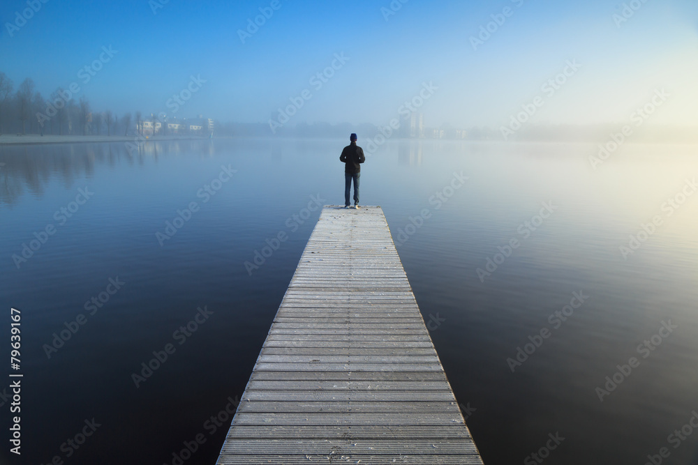 Man standing alone on a jetty, looking over a foggy lake. - obrazy, fototapety, plakaty 