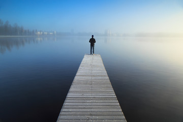 man standing alone on a jetty, looking over a foggy lake.