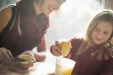 A Woman And A Girl Sitting At A Table, Girl Squeezing The Juice From A Lemon Into A Glass.