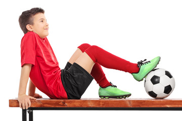 Wall Mural - Relaxed young boy in football uniform sitting on a bench and thi