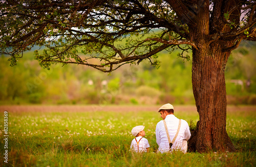 Fotografija  father and son sitting under the tree on spring lawn