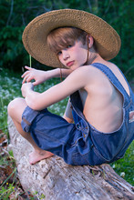 Young Boy Wearing Overalls Sitting On Tree Wearing Straw Hat