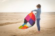© RooM The Agency - Side view of a barefoot boy in jeans standing on a sandy beach shaking a multi coloured patchwork blanket