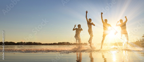 Fotomural Glückliche junge Menschen laufen und springen am See beim Sonnenuntergang