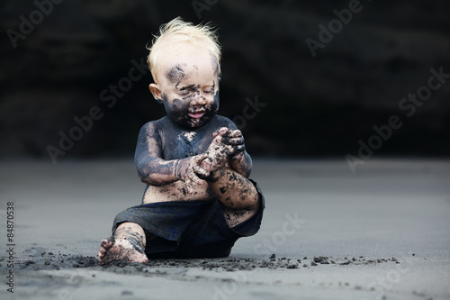 Fotografía Funny portrait of smiling child with dirty face sitting and playing with fun on black sand sea beach before swimming in ocean
