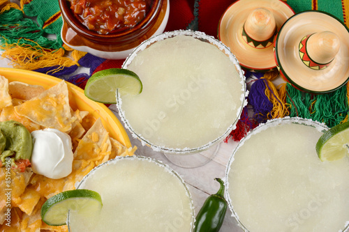 Fotografie, Obraz Margaritas: High angle view of three margarita cocktails surrounded by nachos, chips and salsa on a bright Mexican, table cloth