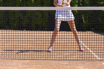 Wall Mural - Slim legs of female tennis athlete behind fishnet barrier.
Young woman stays in ready position close to tennis playground barrier ready to return the ball white dress miniskirt focus on legs