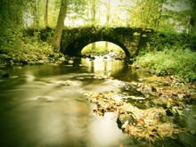 Old Stony Bridge Above Autumn River. Water Of Stream Full Of Colorful Leaves, Leaves On Gravel, Blue Blurred Water Is Running Over Boulders
