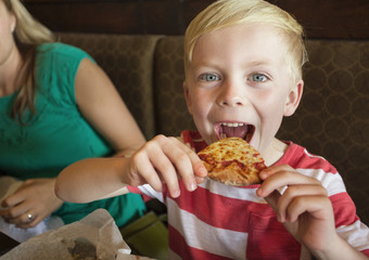 Cute little boy taking a big bite of cheese pizza at a restaurant