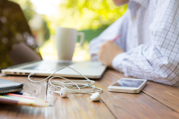 Remote office concept. Business style dressed man sitting at natural country style wooden desk with electronic gadgets around working on laptop drinking coffee sunlight and green terrace on background