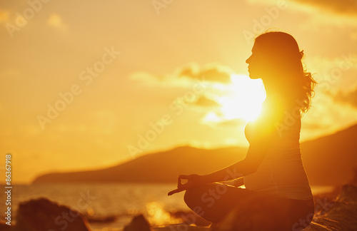 Fotografia Pregnant woman practicing yoga in lotus position on beach at sun