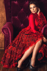 Wall Mural - Portrait of amazing beautiful sexy brunette woman with lovely plump lips in red tango dress posing sitting in a velvety armchair and dreaming. Dark brown luxury velvet background with copyspace