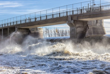 Horizontal Image Of A Concrete Bridge Over A Water Dam With Water Running So Fast Over The Turbine It Churns Violently And Splashes Down The River In The Summer Time.