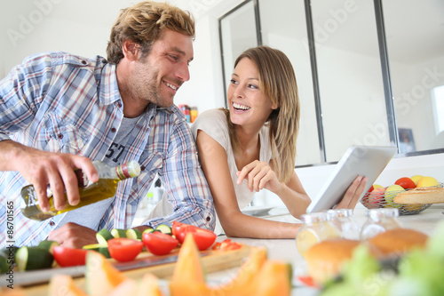 Fotografie, Obraz Cheerful couple in kitchen cooking dinner, using tablet