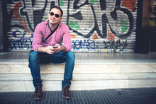 Portrait Of Handsome Man With Phone In Hand, Casually Dressed With Shirt, Jeans And Sunglasses Against Graffiti Painted Wall. Stylish European Man Travelling And Taking Pictures With Mobile Phone