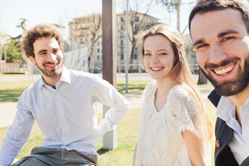 In a sunny spring, a group of three young friends, two men and a woman are sitting on a park bench talking and joking