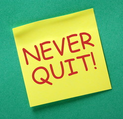 The motivational phrase Never Quit in red text on a yellow sticky note posted on a green notice board asa reminder