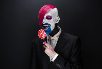 Wall Mural - Clown and Halloween theme: Scary clown with pink hair in a black jacket with candy in hand on a dark background in the studio