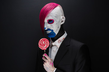 Wall Mural - Clown and Halloween theme: Scary clown with pink hair in a black jacket with candy in hand on a dark background in the studio