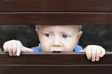 Portrait of abandoned by parents little baby boy with staring blue eyes, sad and lonely face expression, looking out through the fence. Social family problems and children stress and negative emotions