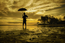 Image Silhouette A Boy Standing And Look At The Back While Holding An Umbrella. Sand And Tree Reflection On Water