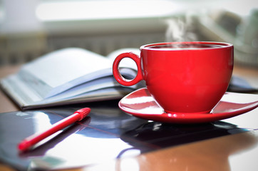 Red cup of hot coffee, notepad and pencil on the desktop in the office. .Focus on the cup.