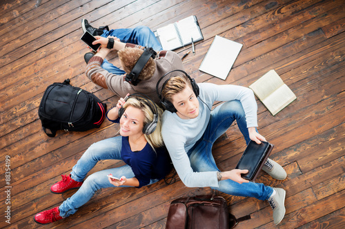 Fotografía Group of students using smartphones and tablet in headphones listening to the music and leaning on each other on wooden floor having notebooks and bags around them