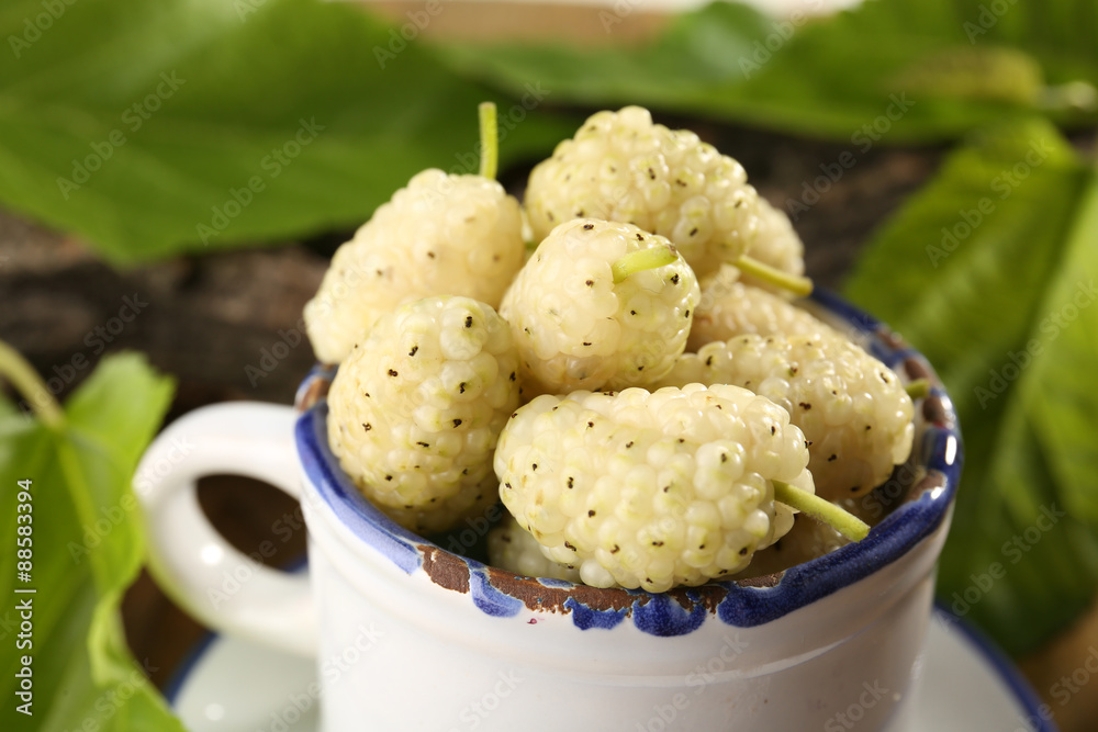 Ripe mulberries in mug with green leaves on table close up
