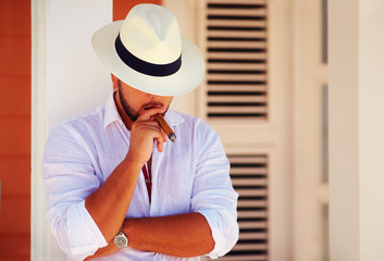 confident handsome man smoking cigar, while leaning on the wall