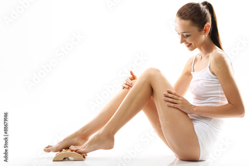 Plakat Masaż stóp. Kobieta w białej bieliźnie masuje stopy drewnianym masażerem 