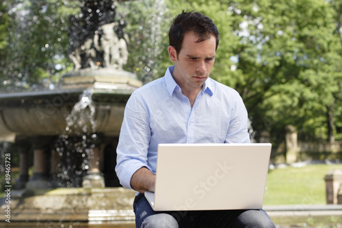Fotografie, Obraz Man sitting a fountain working on laptop computer.