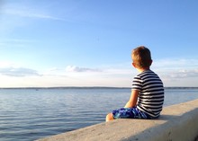 Child Sitting On The Embankment Looking At Water In Summer