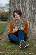 Beautiful young woman sitting next to a tree, holding a book and smiling
