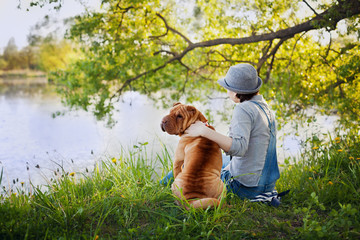 young woman in a hat with dog Shar Pei sitting in the field and looking to the river in golden sunset light, true friends forever, people concept, meditation and relaxation, back to camera