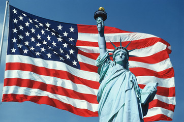 This is an American flag on a flagpole waving in the wind set against a blue sky. The top half of the Statue of Liberty is digitally composited onto the right side of the flag with the torch coming up in the blue sky. This is a digitally created image.