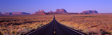 This Is Route 163 That Runs Through The Navajo Indian Reservation. The Road Runs Up The Middle And Gets Smaller Into Infinity. The Red Rocks Of Monument Valley Are In The Background. The Scrub Plants Of The Desert Are On Either Side Of The Road.