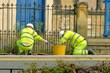 Two council workers cleaning drains outside iron church gates.