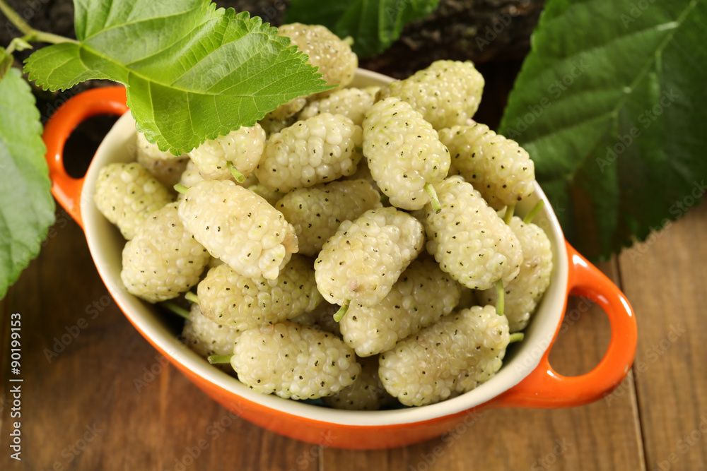 Ripe mulberries in bowl with green leaves on table close up