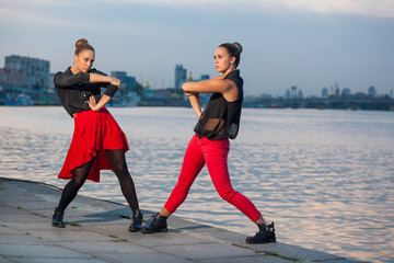 Two young beautiful twin sisters are dancing waacking dance in the city background near river. showing the different style and pose of modern dance with black and red dress near water on summer time.