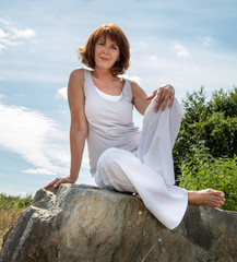 senior zen - smiling, beautifully aging woman sitting on a stone for outdoors yoga session wearing white seeking serenity and wellness in a park,summer daylight