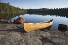 Yellow Canoe On Rocky Shore Of Calm Lake With Pine Trees