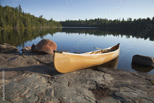 Cuadros en Lienzo Yellow canoe on rocky shore of calm lake with pine trees
