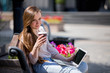 Young beautiful woman sitting on a bench with laptop and coffee in hand
