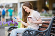 Young beautiful woman sitting on a bench on the street with coffee cup in hand and shopping bags. Drinking coffee, smiling and using digital tablet.
