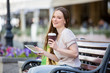 Young beautiful woman sitting on a bench on the street with coffee cup in hand and shopping bags. Drinking coffee, smiling and using digital tablet.
