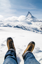 Men's Legs In The Trekking Boots Lying On The Snow With The Background Of Matterhorn, Switzerland.