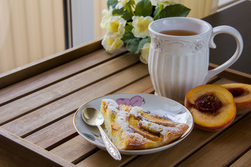 piece of peach pie on a white plate, white mug with tea on a wooden surface and yellow flowers in the background, natural light from the window. Horizontal photo