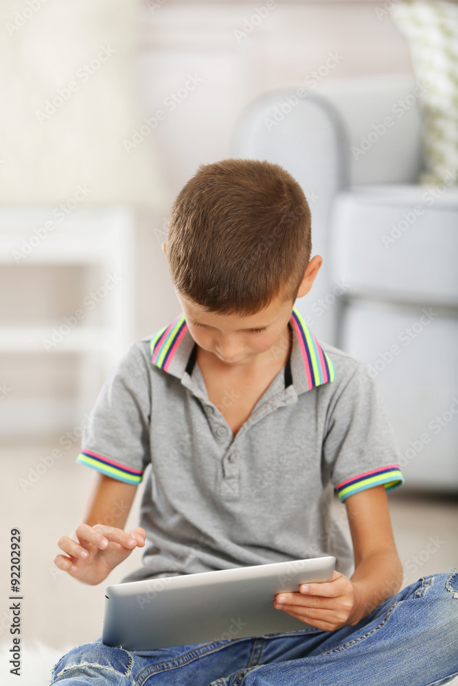 Little boy with digital tablet sitting on carpet, on home interior background